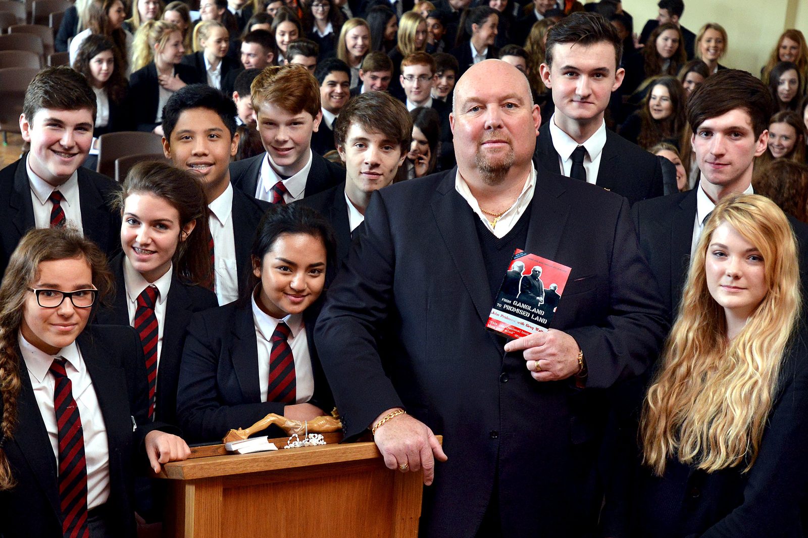John Pridmore standing in a school hall surrounded by secondary students, holding a copy of his book